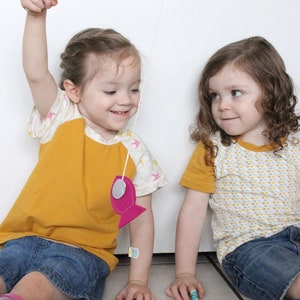 May include: Two children play a fishing game with a wooden rod and string. One child wears a yellow and white shirt with a pink fish attached to the string. The other child wears a yellow shirt with a white and yellow geometric pattern.