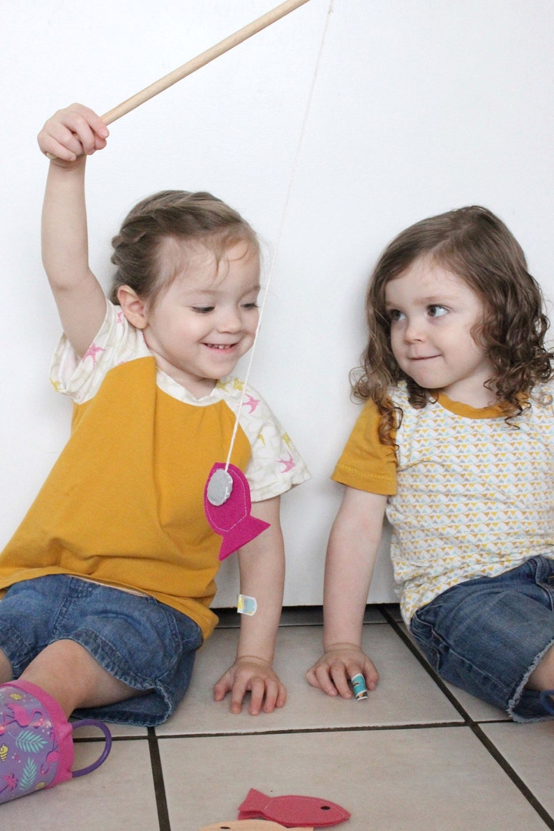 May include: Two children play a fishing game with a wooden rod and string. One child wears a yellow and white shirt with a pink fish attached to the string. The other child wears a yellow shirt with a white and yellow geometric pattern.