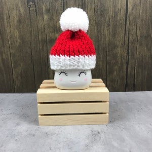 May include: A white ceramic mug with a smiling face, wearing a small red and white knit Santa hat with a white pom-pom. The mug is sitting on a small wooden crate, against a wood-grain background.