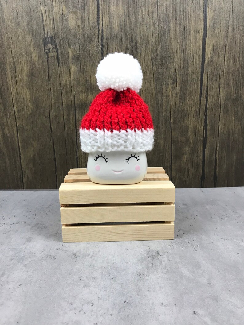 May include: A white ceramic mug with a smiling face, wearing a small red and white knit Santa hat with a white pom-pom. The mug is sitting on a small wooden crate, against a wood-grain background.