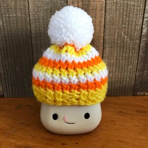 Puede incluir: Una taza de cer&aacute;mica blanca con una cara sonriente que lleva un gorro de punto a rayas amarillo, naranja y blanco con un pomp&oacute;n blanco.
