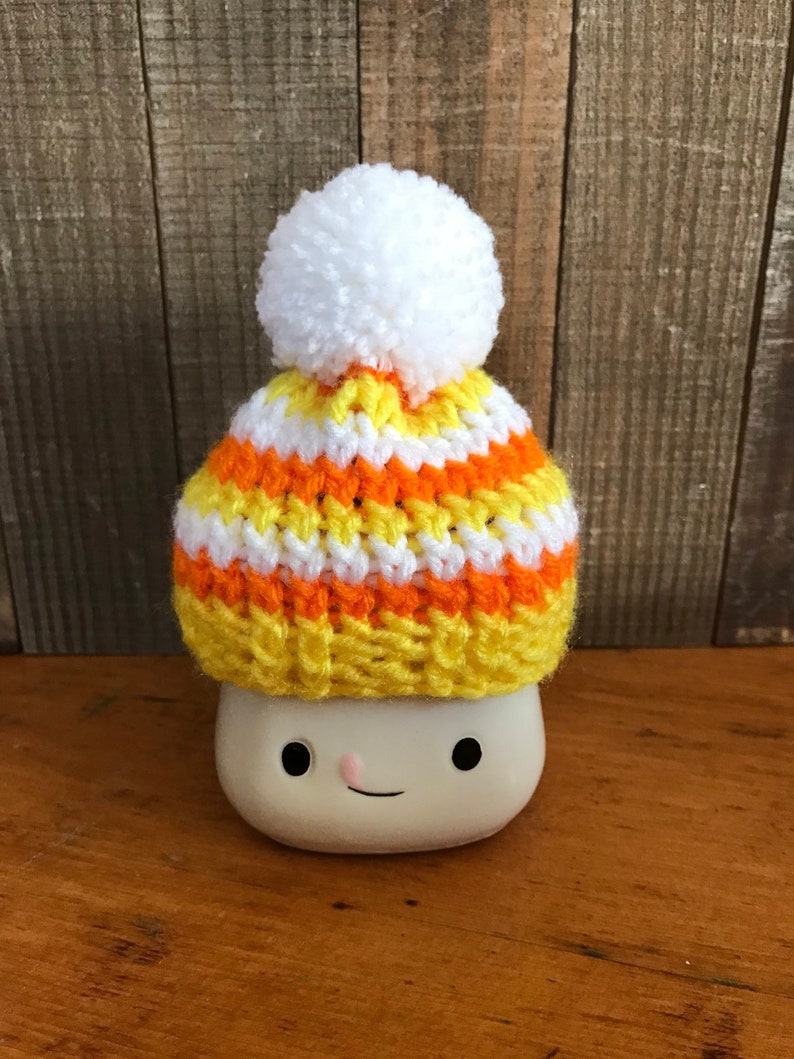 Puede incluir: Una taza de cer&aacute;mica blanca con una cara sonriente que lleva un gorro de punto a rayas amarillo, naranja y blanco con un pomp&oacute;n blanco.