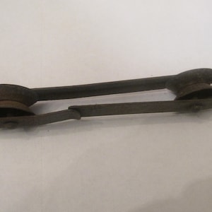 Może przedstawiać: Rustykalne metalowe narzędzie z dwoma kołami na obu końcach. Narzędzie jest przeznaczone do ciągnięcia lub dokręcania czegoś.