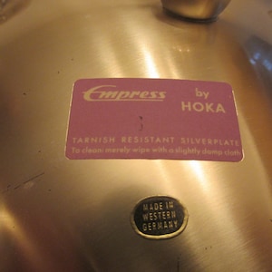 May include: A gold-colored metal serving dish with a label that reads "Empress by Hoka Tarnish Resistant Silverplate To clean merely wipe with a slightly damp cloth" and a smaller label that reads "Made in Western Germany".