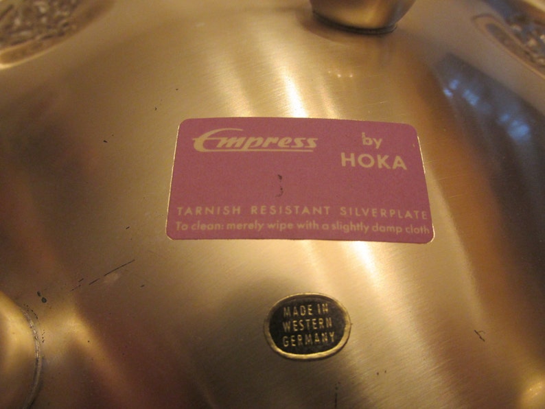 May include: A gold-colored metal serving dish with a label that reads "Empress by Hoka Tarnish Resistant Silverplate To clean merely wipe with a slightly damp cloth" and a smaller label that reads "Made in Western Germany".