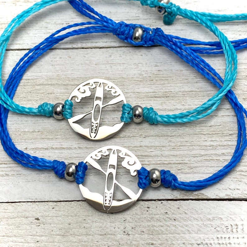 May include: Two blue and turquoise adjustable bracelets with silver charms featuring a kayak silhouette against a wave background.