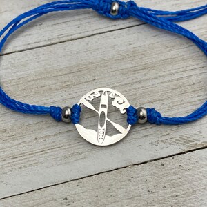 May include: A blue cord bracelet with a silver charm featuring a cutout design of a kayak on a lake with clouds.