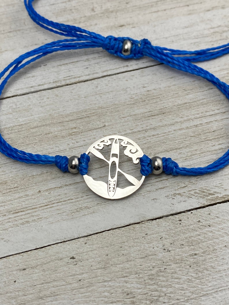 May include: A blue cord bracelet with a silver charm featuring a cutout design of a kayak on a lake with clouds.