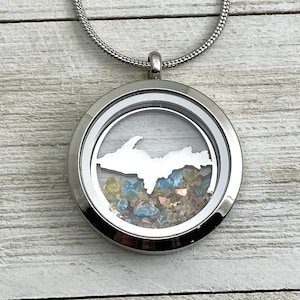 Può includere: Una collana con un ciondolo d'argento e un inserto in vetro trasparente che presenta un ritaglio della Upper Peninsula del Michigan. Il ciondolo è riempito di sabbia colorata e piccole pietre.