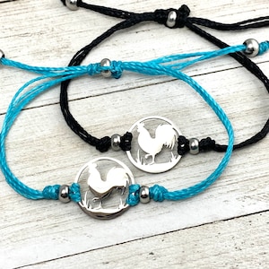 Può includere: Due braccialetti dell'amicizia con ciondoli a forma di gallo argentati. Un braccialetto è in cordoncino nero con perline argentate e l'altro in cordoncino turchese con perline argentate.
