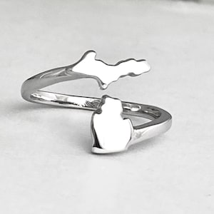Puede incluir: Un anillo plateado con la forma del estado de Michigan. El anillo tiene un diseño abierto, con las penínsulas superior e inferior de Michigan como punto focal. El anillo está hecho de un metal brillante y reflectante.