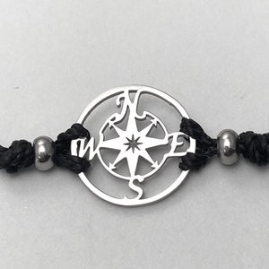 May include: A black cord bracelet with a silver compass charm. The compass features a star in the center and the letters N, S, E, and W around the edge.