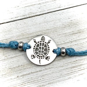 May include: A silver charm bracelet with a cutout turtle design. The bracelet is made with blue cord and silver beads.