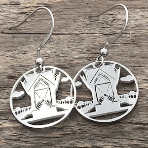 Puede incluir: Un par de pendientes de plata con un diseño recortado de un granero con nubes y un campo. Los pendientes están sobre una superficie de madera.