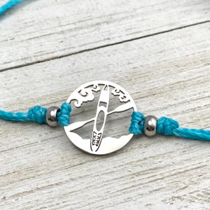 May include: A turquoise blue cord bracelet with a silver charm featuring a silhouette of a kayak paddling through waves.