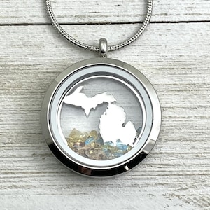 May include: A silver locket necklace with a clear glass insert featuring a white outline of the state of Michigan and colorful sand inside.
