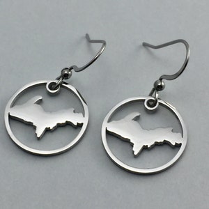 May include: A pair of silver-colored dangle earrings. Each earring features a circular frame with a cutout of the Upper Peninsula of Michigan. The earrings have a hook closure.