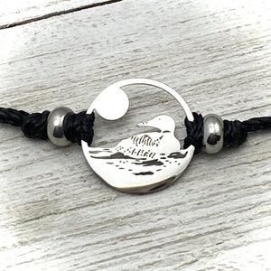 Könnte beinhalten: Ein silbernes Armband mit einem runden Anhänger, der die Silhouette eines Seetaucher auf einem See zeigt. Der Anhänger ist an einem schwarzen Band mit silbernen Perlen befestigt.