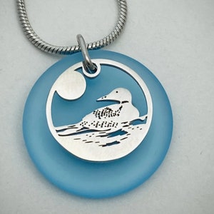 May include: A silver pendant necklace with a blue glass circle featuring a cut-out design of a loon on a lake. The loon is facing left and the moon is visible in the background.