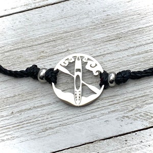 May include: A silver bracelet with a round charm featuring a silhouette of a kayak with paddles and waves. The bracelet is made of black cord and has silver beads.