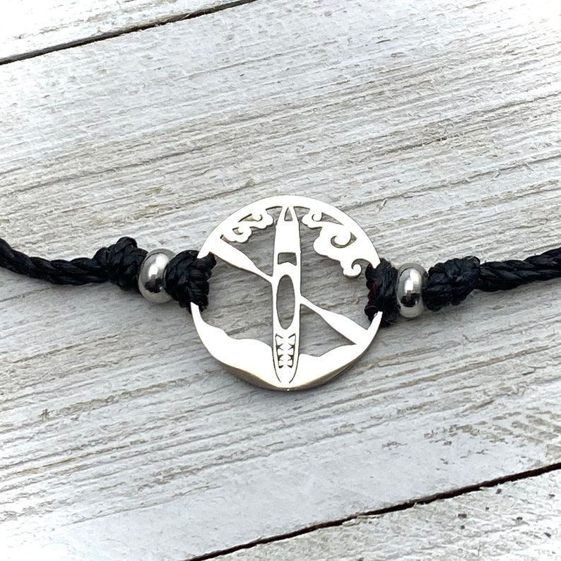 May include: A silver bracelet with a round charm featuring a silhouette of a kayak with paddles and waves. The bracelet is made of black cord and has silver beads.
