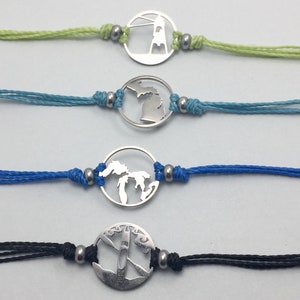 Op de afbeelding: Vier verschillende armbandjes met zilveren bedels. De bedels zijn een vuurtoren, de staat Michigan, de Grote Meren en een kajak. De armbandjes zijn op een witte achtergrond.