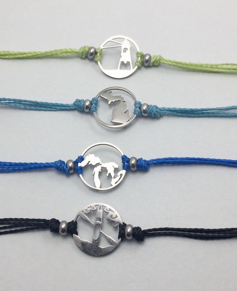 Op de afbeelding: Vier verschillende armbandjes met zilveren bedels. De bedels zijn een vuurtoren, de staat Michigan, de Grote Meren en een kajak. De armbandjes zijn op een witte achtergrond.