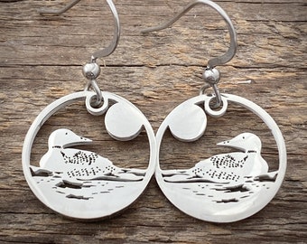 Puede incluir: Un par de pendientes de plata con un diseño de silueta de loón. Los pendientes son circulares y tienen un diseño recortado de un loón nadando en un lago con una luna en el fondo.