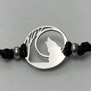 Op de afbeelding: Een zilveren armband met een ronde bedel met een wolf die naar de maan huilt. De bedel is bevestigd aan een zwart koord met zilveren kralen.