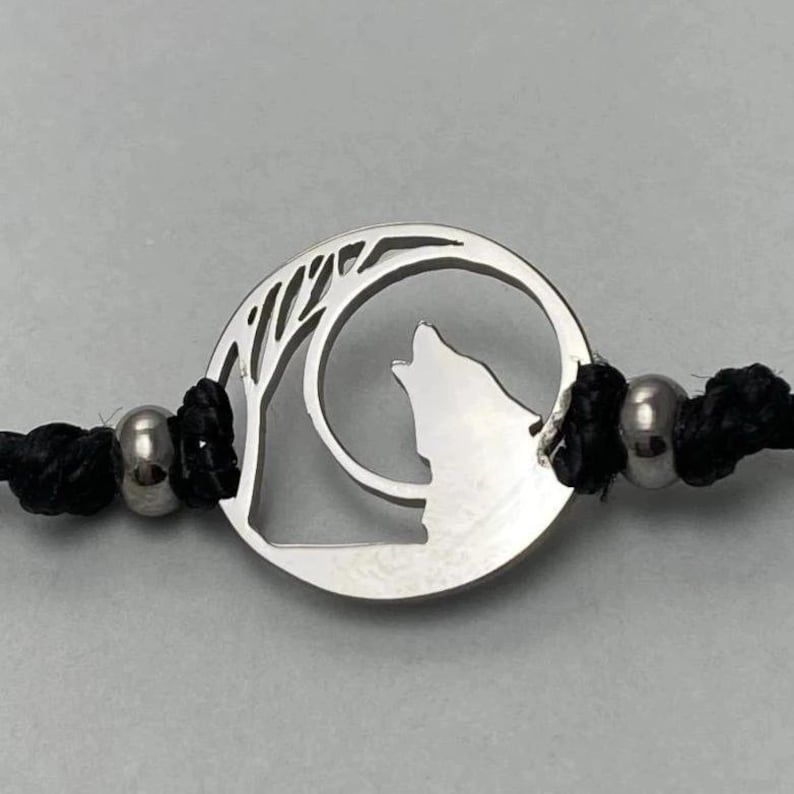 Op de afbeelding: Een zilveren armband met een ronde bedel met een wolf die naar de maan huilt. De bedel is bevestigd aan een zwart koord met zilveren kralen.