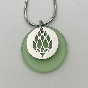 May include: A green glass pendant necklace with a silver metal charm featuring a cut-out design of a hop cone.