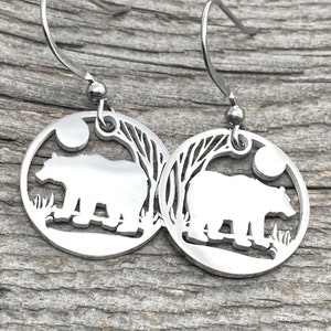 May include: A pair of silver earrings with a bear silhouette design. Each earring features a circular pendant with a bear standing in front of a tree and a moon.