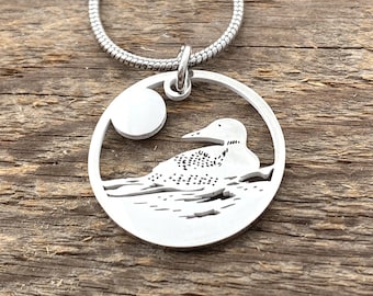 May include: A silver necklace with a round pendant featuring a loon silhouette on a lake with a moon in the background.