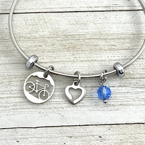 May include: A silver bangle bracelet with three charms: a bike, a heart, and a blue crystal.