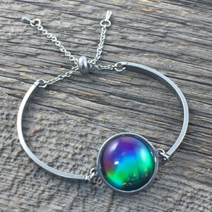 May include: A silver bracelet with a round, iridescent, multi-colored glass cabochon. The bracelet has a sliding clasp.