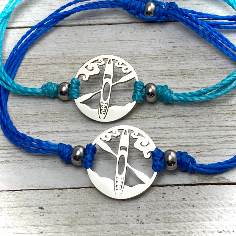 May include: Two blue and turquoise braided cord bracelets with silver charms featuring a kayak silhouette inside a circle with waves.
