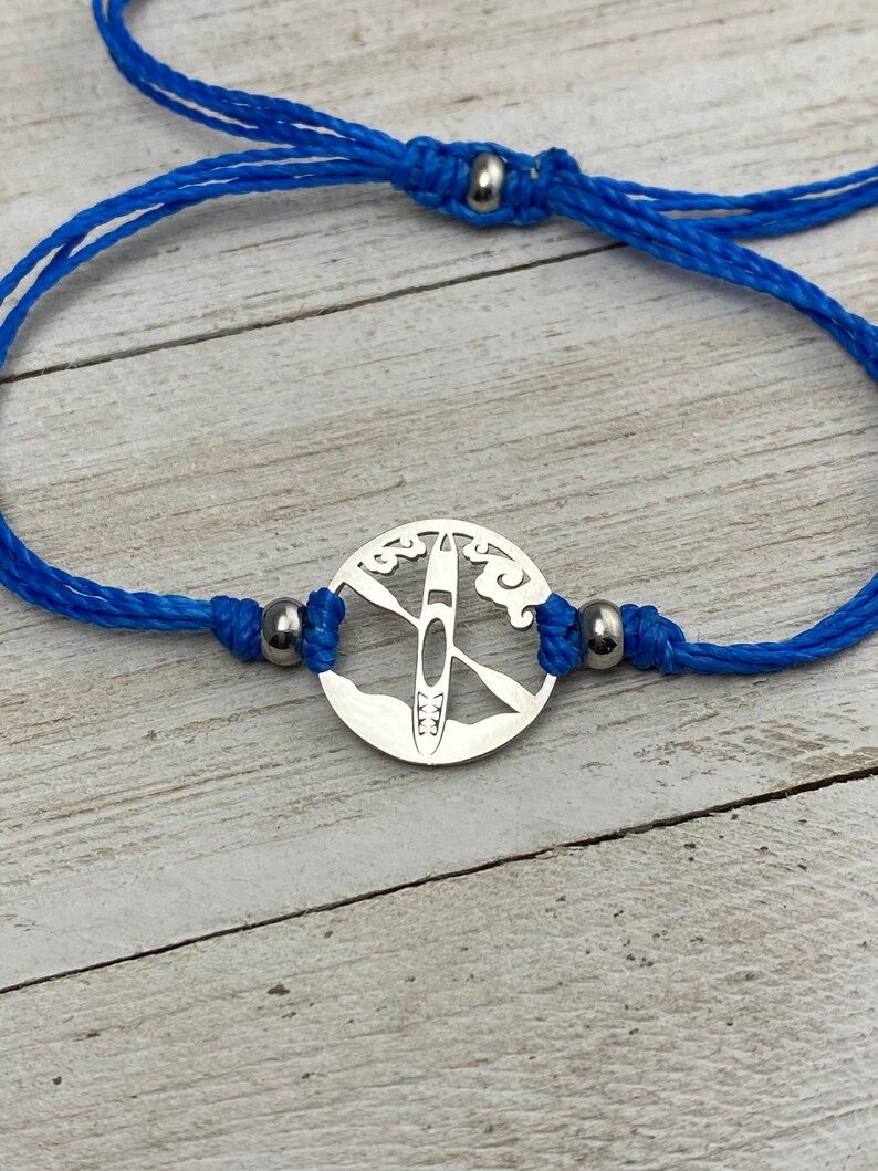 May include: A blue cord bracelet with a silver charm featuring a cutout design of a kayak on a lake with clouds.