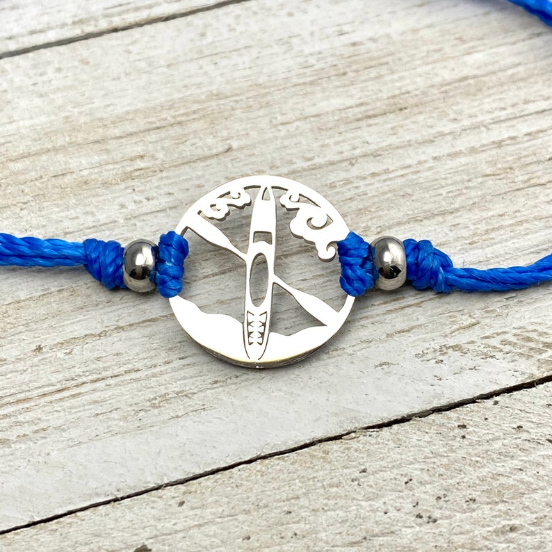 May include: A blue cord bracelet with silver metal beads and a silver charm featuring a silhouette of a kayak with paddles and waves.