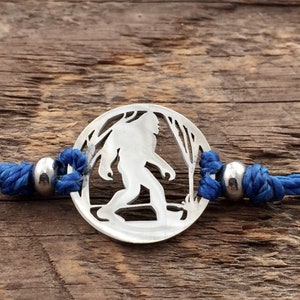 Puede incluir: Un brazalete de cordón azul con un dije de plata que presenta una silueta de Bigfoot caminando por el bosque.