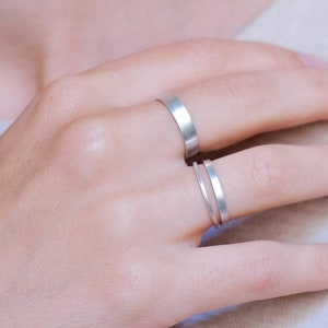 Op de afbeelding: Twee zilveren ringen aan een vinger. De bovenste ring is een eenvoudige band, terwijl de onderste ring een dunne band is met een iets bredere band eromheen gewikkeld.