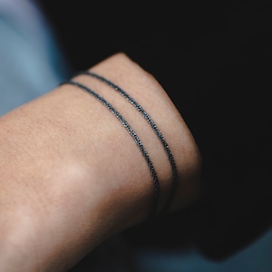 Puede incluir: Dos pulseras finas de cadena negra apiladas en la muñeca de una persona.