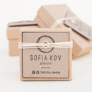 May include: A small, brown cardboard box with a white string tied around it. The box has a label that says "Sofia Kov Jewelry" and a circle with a gold ring in the center. The box is sitting on a white surface.