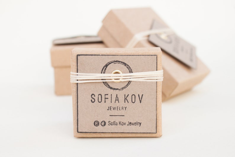 May include: A small, brown cardboard box with a white string tied around it. The box has a label that says "Sofia Kov Jewelry" and a circle with a gold ring in the center. The box is sitting on a white surface.