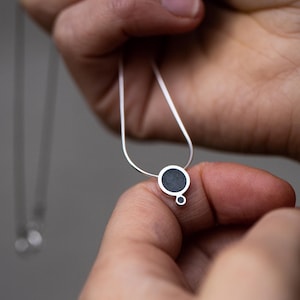 Puede incluir: Un collar de plata con un colgante circular negro. El colgante tiene un c&iacute;rculo m&aacute;s peque&ntilde;o unido a la parte inferior.