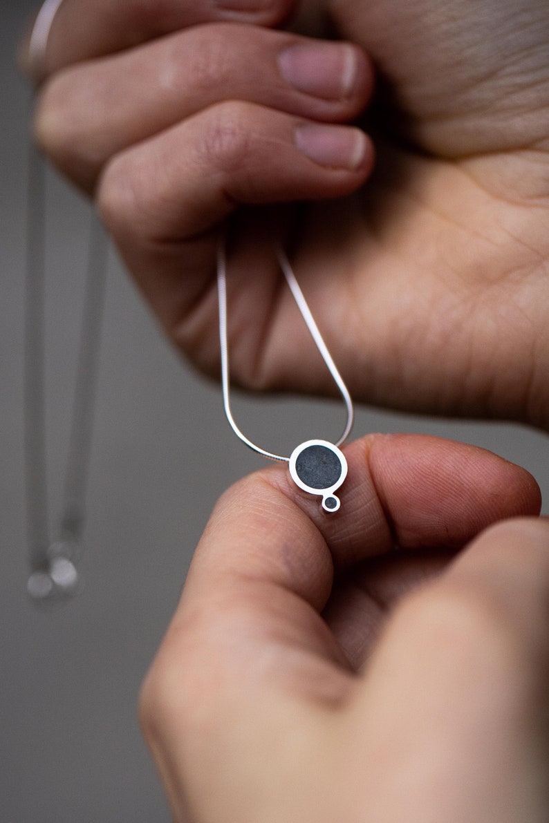 Puede incluir: Un collar de plata con un colgante circular negro. El colgante tiene un c&iacute;rculo m&aacute;s peque&ntilde;o unido a la parte inferior.