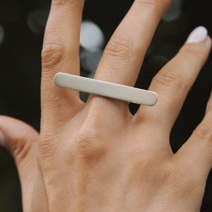 Könnte beinhalten: Ein silberfarbener Ring mit einem langen, horizontalen, rechteckigen Balkendesign, das sich über zwei Finger erstreckt. Die Metalloberfläche hat ein glattes, mattes Finish mit abgerundeten Kanten, was ein klares und einheitliches Erscheinungsbild erzeugt.