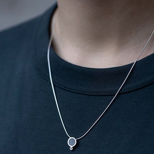 Puede incluir: Un collar de cadena de plata con un peque&ntilde;o colgante redondo. El colgante es negro y tiene un peque&ntilde;o c&iacute;rculo plateado unido a la parte inferior.