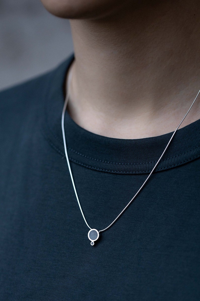 Puede incluir: Un collar de cadena de plata con un peque&ntilde;o colgante redondo. El colgante es negro y tiene un peque&ntilde;o c&iacute;rculo plateado unido a la parte inferior.