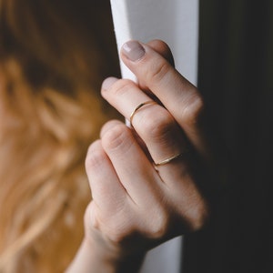 May include: A close-up of a hand wearing two thin gold rings. The hand is reaching towards a white curtain.