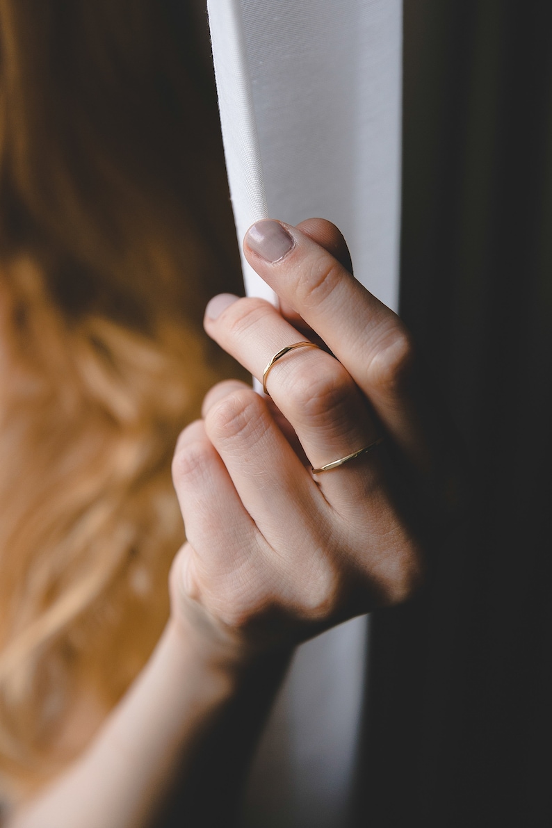 May include: A close-up of a hand wearing two thin gold rings. The hand is reaching towards a white curtain.